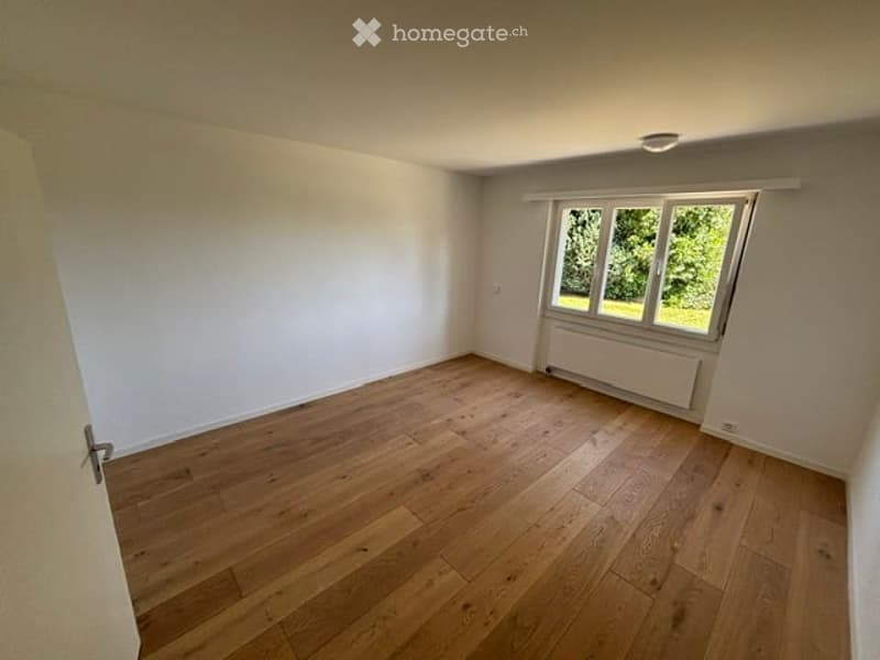 Empty room with wooden flooring, white walls, and a large window with a view of greenery outside.