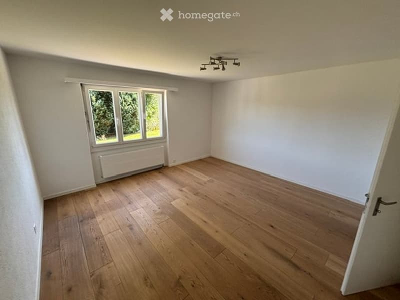 Empty room with wooden flooring, white walls, large window with natural light, radiator, and ceiling light fixture.