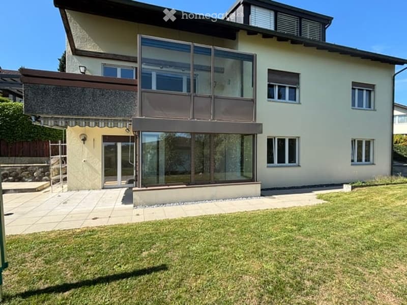 Two-story house with a modern design, large glass balcony, and well-maintained lawn.
