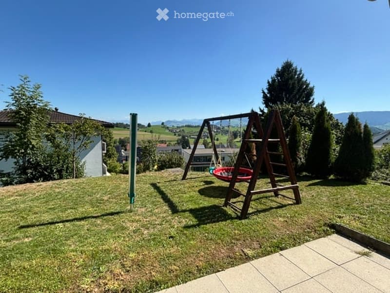 A well-maintained garden with a swing set, grass, trees, and a clear blue sky.