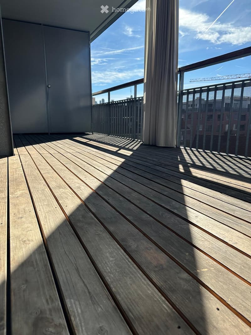 Wooden deck with metal railing, overlooking a cityscape with clear blue sky and scattered clouds.