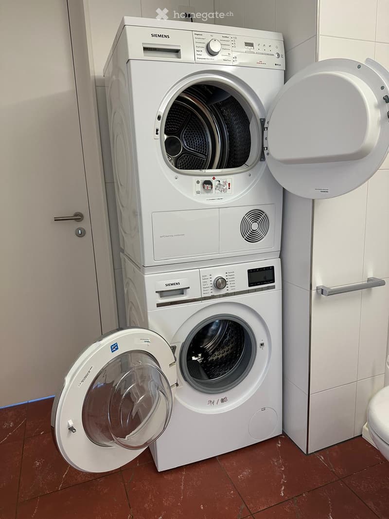 Bathroom with a stacked Siemens washing machine and dryer, both with open doors, placed on a tiled floor.