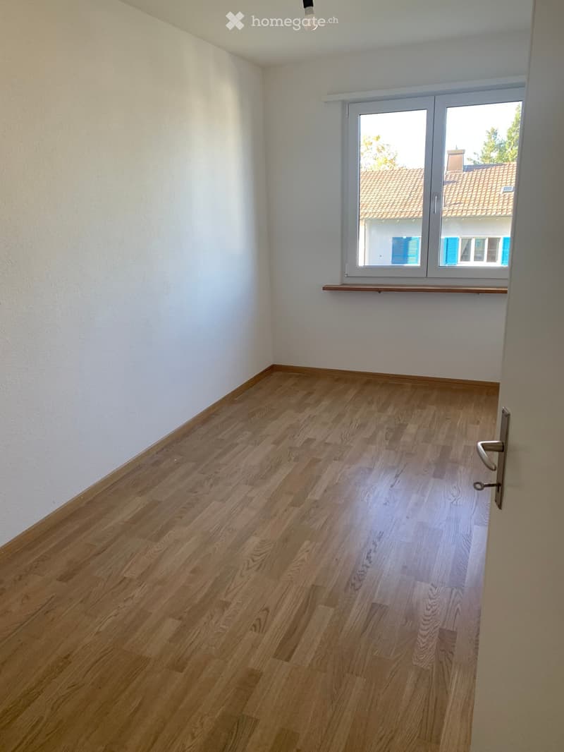 Empty room with light wooden flooring, white walls, and a large window with a view of a neighboring building.
