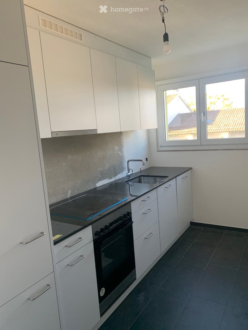 Modern kitchen with white cabinets, black countertops, a sink, and an oven. The space is well-lit with a window offering a view of the outside.