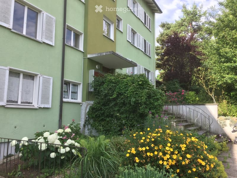 A two-story green building with multiple windows and shutters, surrounded by a garden with colorful flowers and greenery.