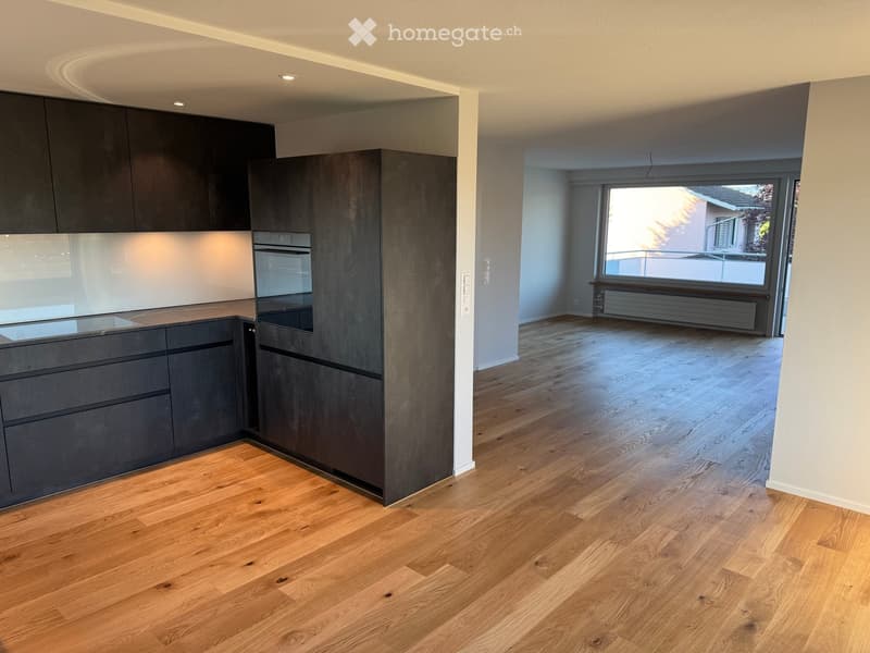 Modern kitchen with dark grey cabinets, a built-in oven, and a sleek countertop, connected to a bright living area with wooden flooring and large windows.
