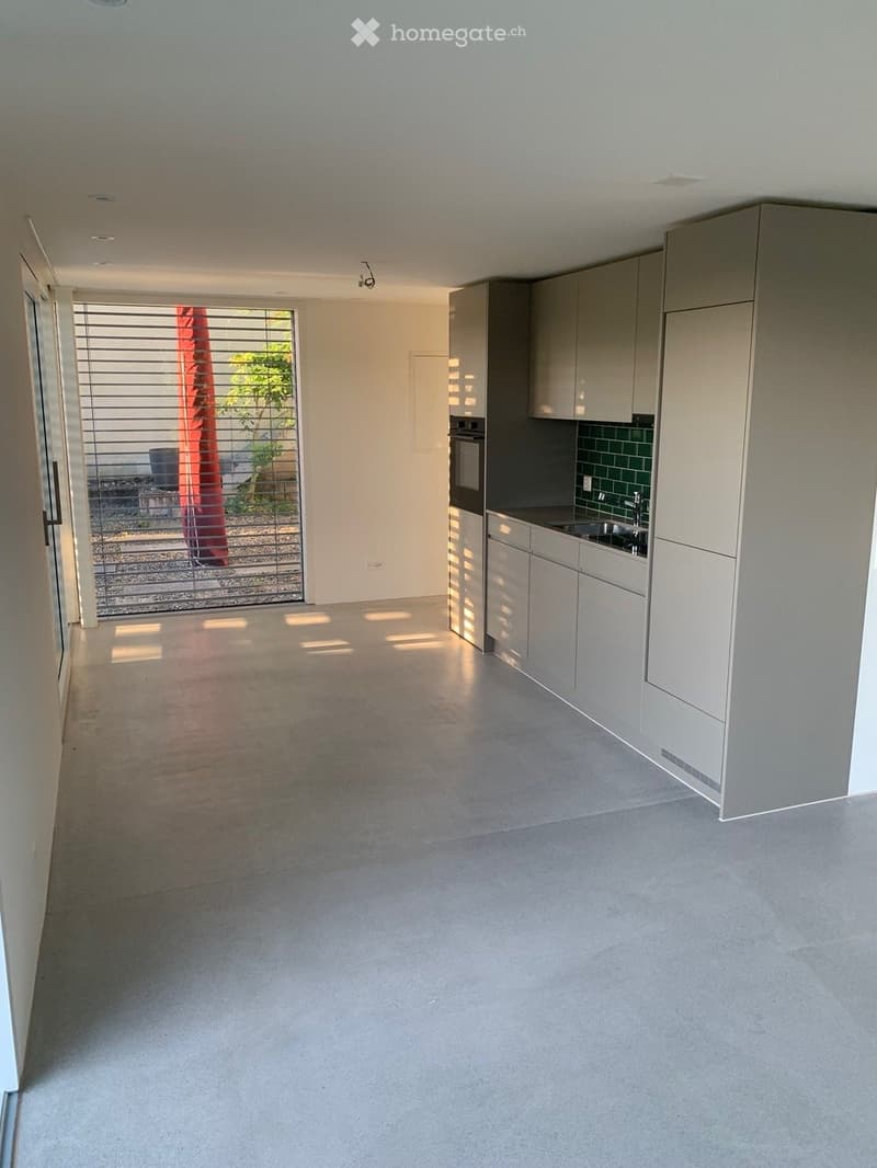 Empty kitchen with modern grey cabinets, stainless steel appliances, and a large window with blinds leading to an outdoor area.