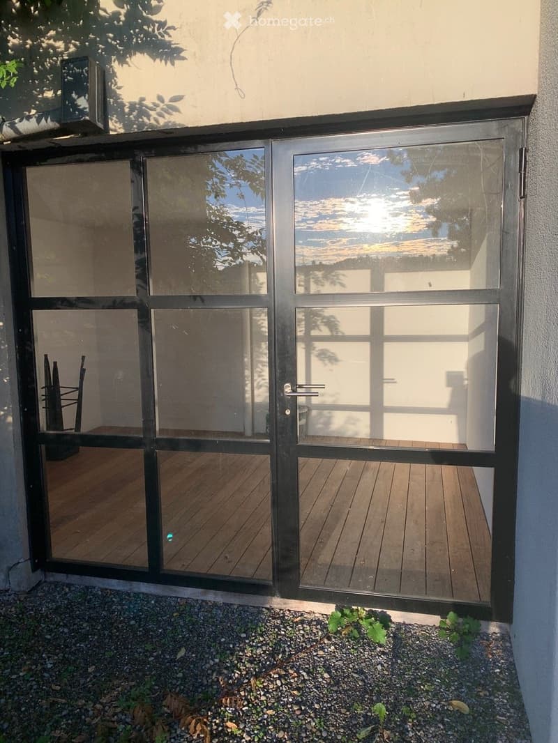 A modern glass balcony door with black frame, leading to a wooden deck area. The deck is visible through the glass panels, with a small garden area in front.