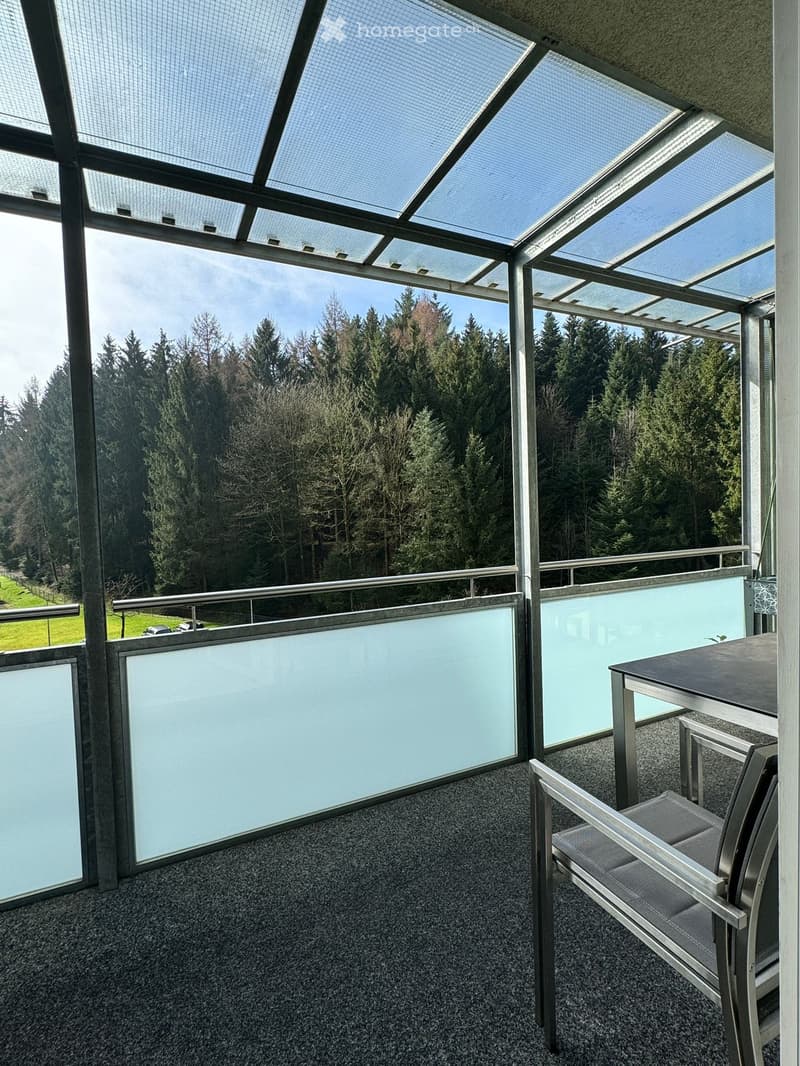 Balcony with a view of a forested area, featuring a table and a chair, with a modern glass ceiling and metal railings.
