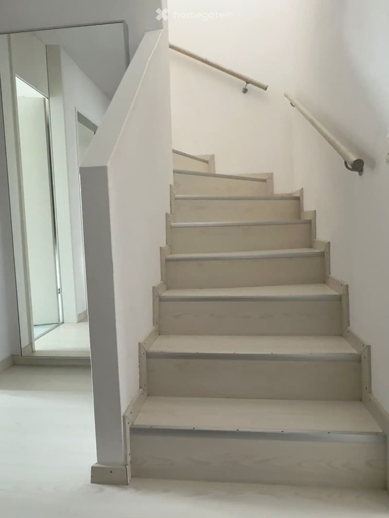 A modern staircase with light-colored wooden steps and white walls, featuring a handrail on one side and a mirror on the adjacent wall.