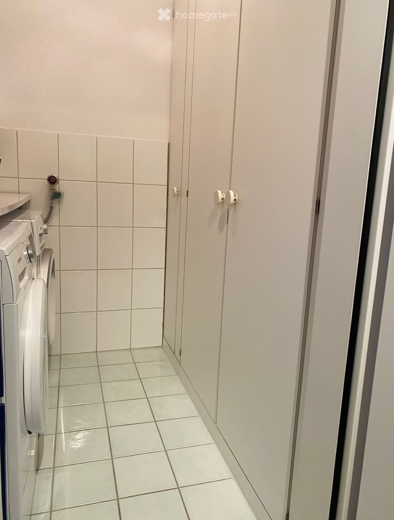 Small bathroom with white tiled walls and floor, featuring a washing machine and a cabinet.