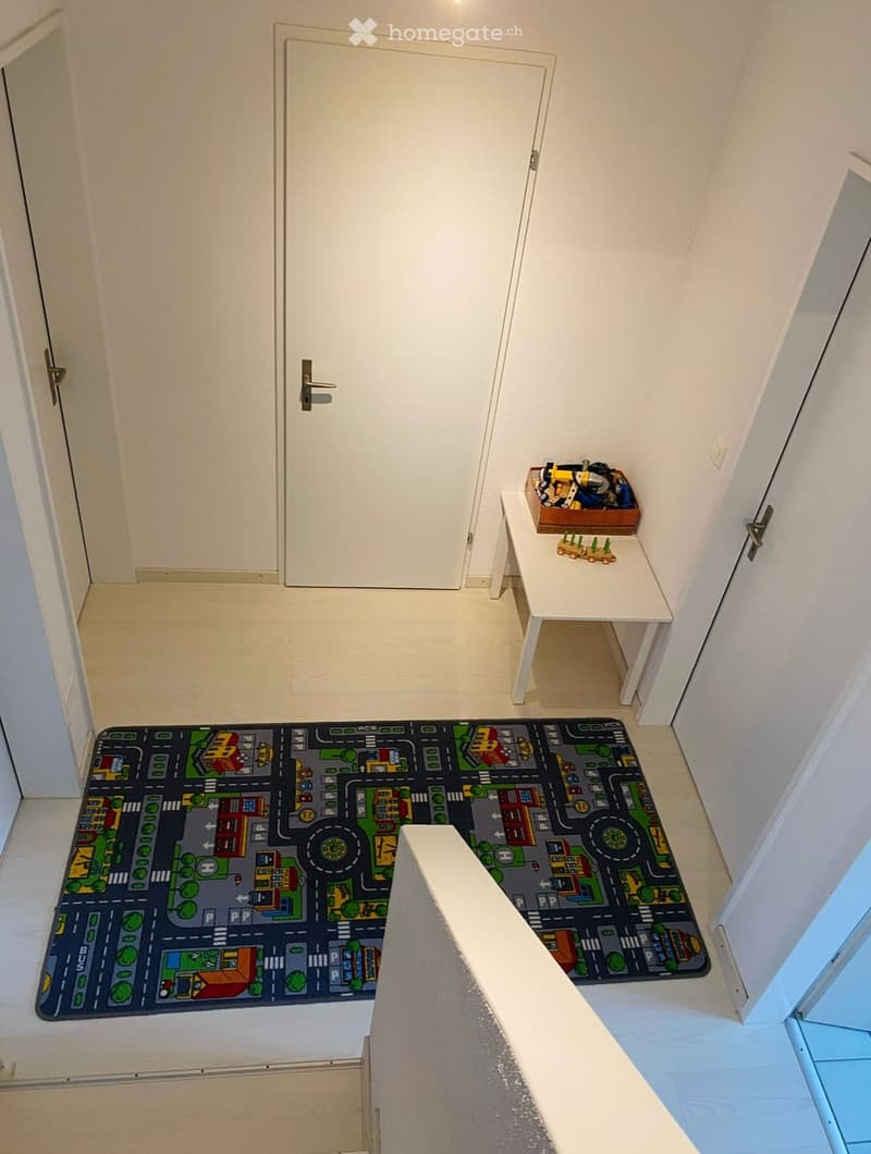 A small, well-lit room with a colorful play mat featuring a road design, a small white table with a toy car and basket, and two doors.