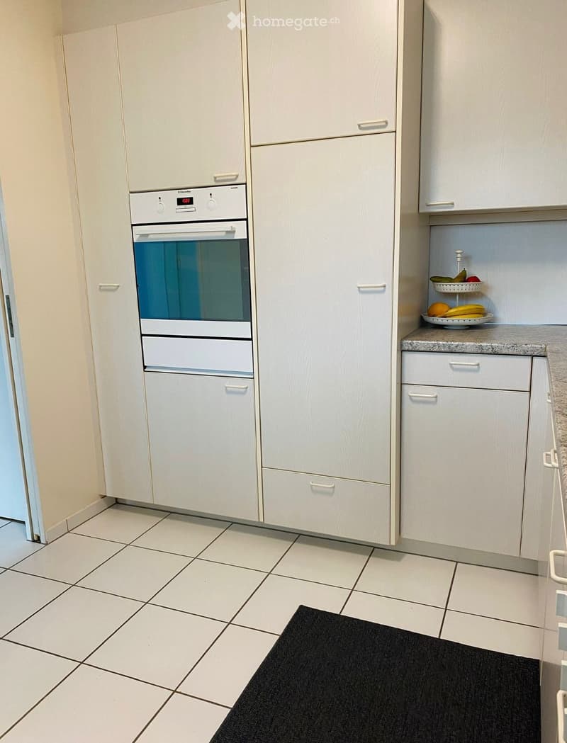 Modern kitchen with white cabinets, a built-in oven, and a countertop with a fruit bowl. The floor is tiled, and there is a black rug in the foreground.