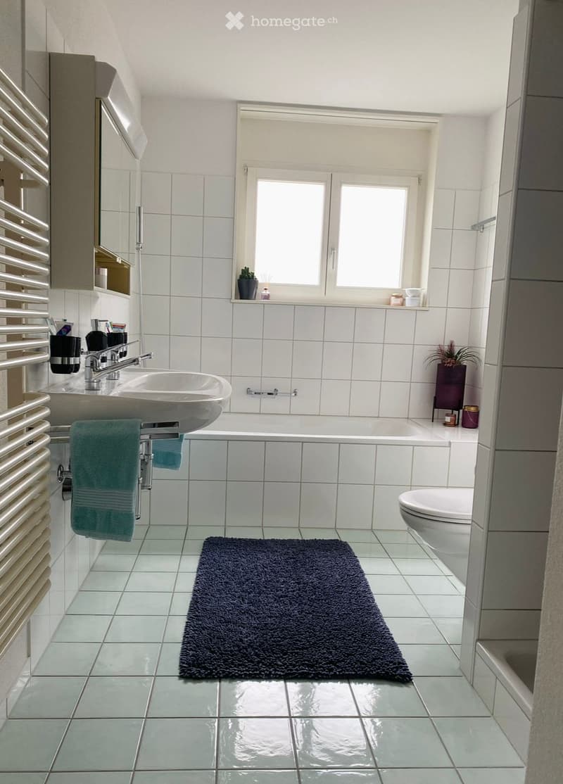 Modern bathroom with white tiles, a double sink, bathtub, toilet, radiator, and a dark blue bath mat on the floor.