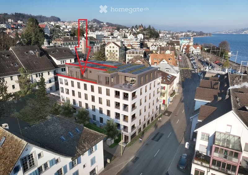 Aerial view of a multi-story residential building with balconies and solar panels on the roof, located in a cityscape with surrounding houses, trees, and a waterfront view.