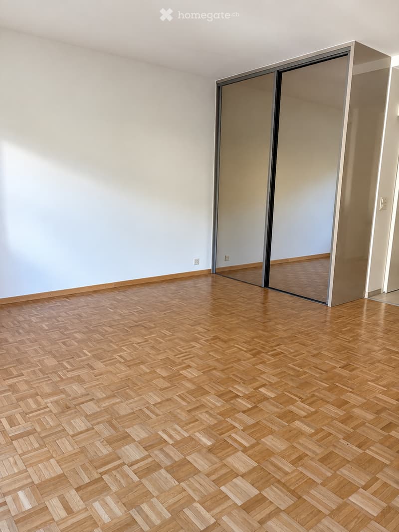Empty room with light-colored walls and a herringbone-patterned wooden floor. A large sliding glass door leads to another room.