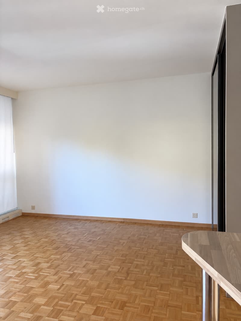 Empty room with light grey walls and herringbone wooden flooring, featuring a large window with blinds and a small wooden table in the corner.