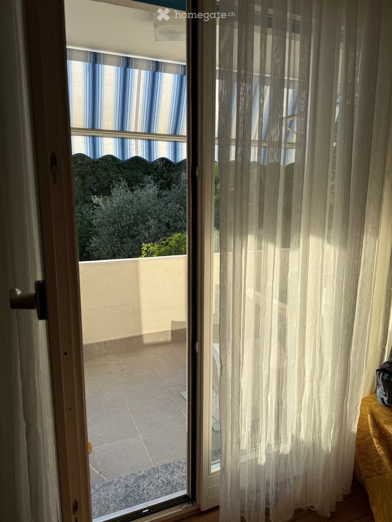 View of a tiled balcony through a large window with sheer white curtains and striped blue and white blinds. The balcony has a tiled floor and is bordered by a low wall with a view of greenery.