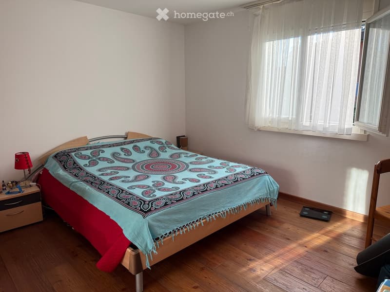 Cozy bedroom with wooden floor, large window with white curtains, bed with colorful patterned bedspread, nightstand with lamp, and a wooden chair.