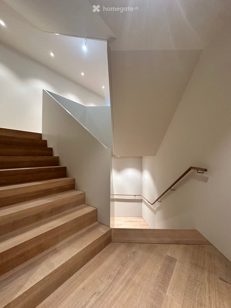 Modern staircase with wooden steps and a metal handrail, leading to an open area with recessed lighting and a view of another room.