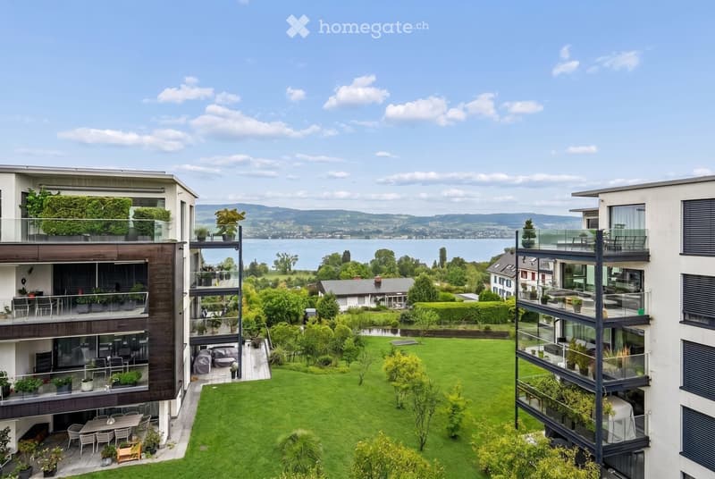 Modern multi-story residential building with balconies, lush green lawn, and scenic lake view in the background.