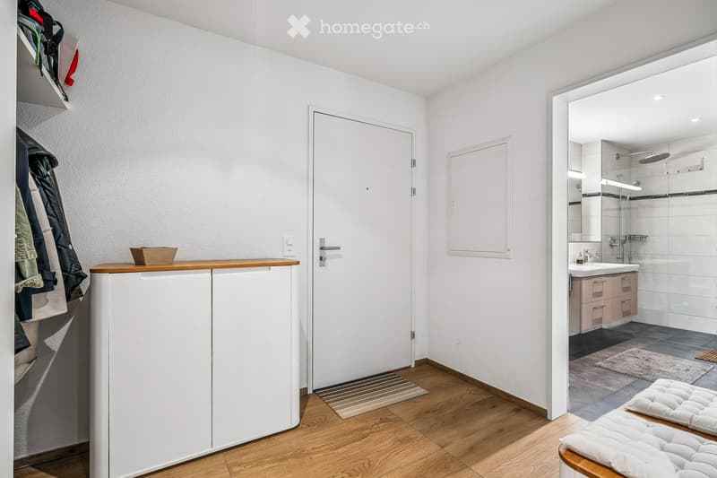 A modern hallway with a wooden bench, a white cabinet, a mirror, and a door leading to a bathroom. The bathroom features a sink, faucet, shower, and a picture frame on the wall.
