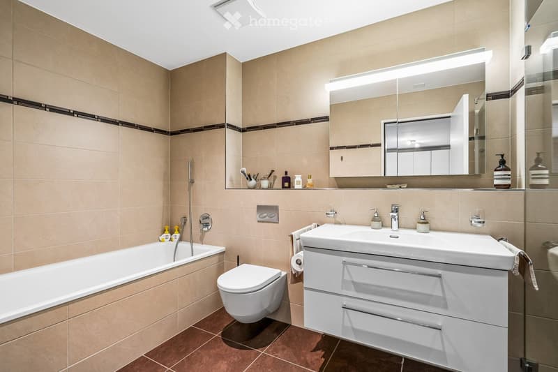 Modern bathroom with beige tiles, a bathtub, toilet, sink with a mirrored cabinet, and various toiletries.
