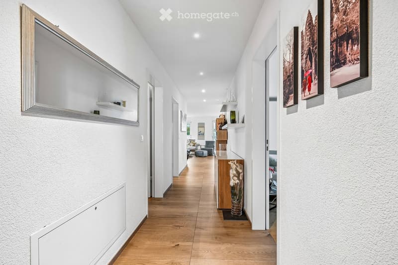 Modern hallway with light wooden flooring, white walls, built-in shelving, and artwork displayed on the walls. The hallway leads to a room with visible furniture, including a sofa and a small table.