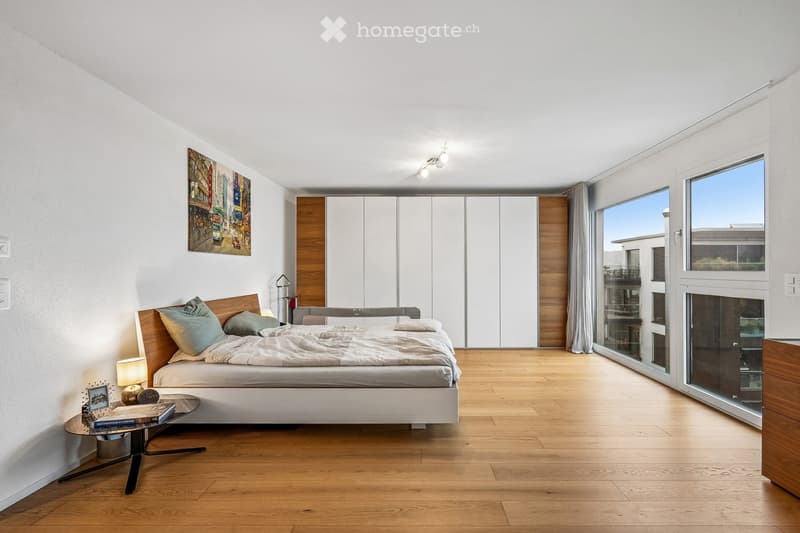 Modern bedroom with wooden flooring, large bed with pillows and blankets, nightstand with lamp, small table with books and decorative items, large windows with view, and built-in wardrobe.