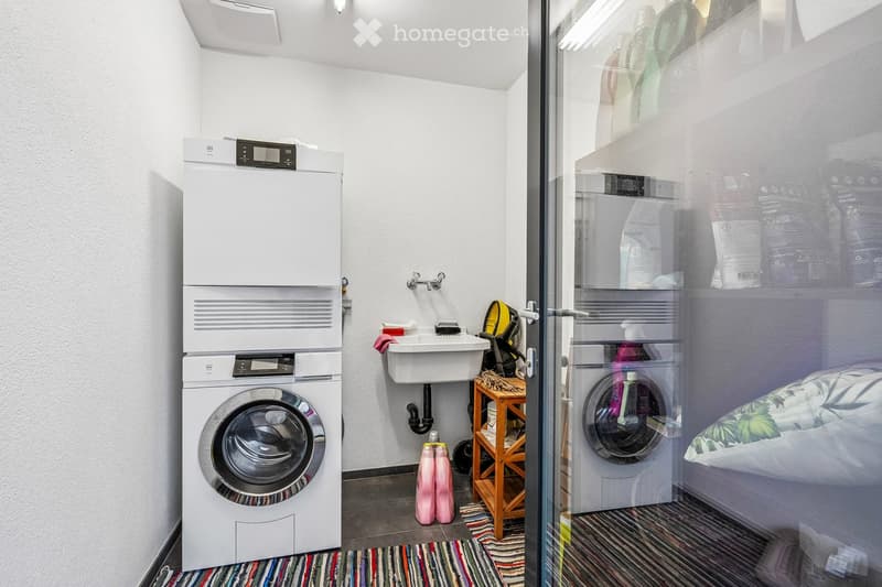 Bathroom with a washing machine, sink, and small wooden stool holding items. The space is well-lit with white walls and a tiled floor.