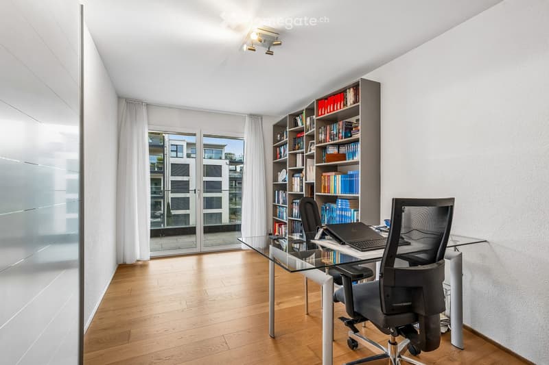 Modern office space with a glass desk, office chairs, a laptop, a bookshelf filled with books, and large windows with white curtains.