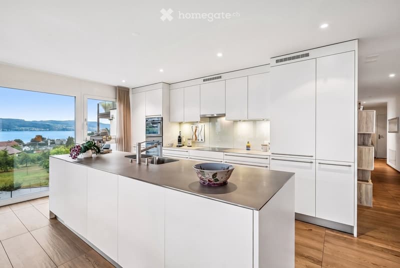 Modern kitchen with white cabinets, stainless steel countertops, and a large window offering a scenic view of a lake and mountains.