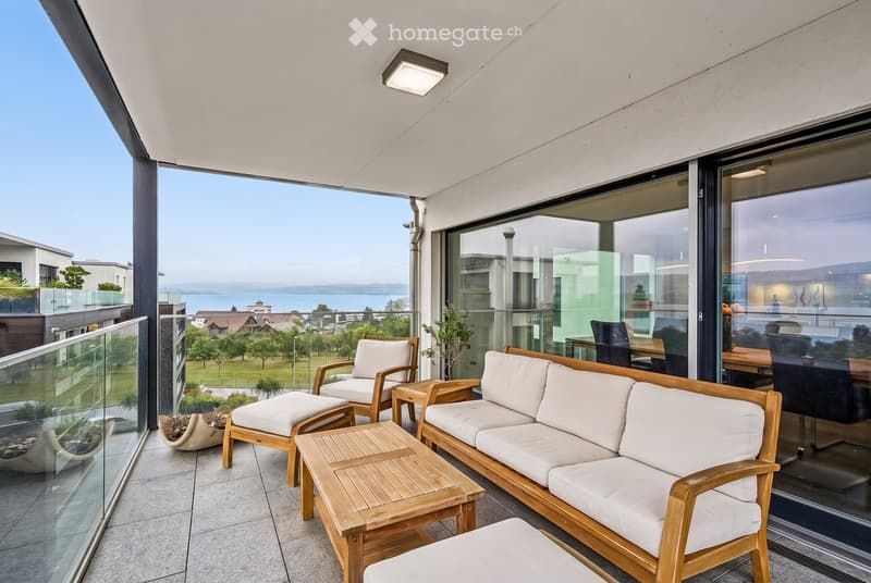 Modern balcony with white cushioned furniture, wooden tables, and a scenic lake view.