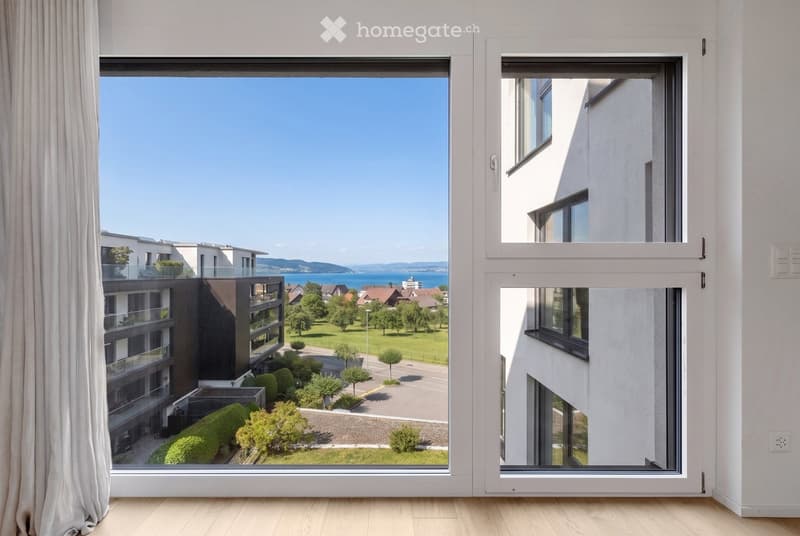 Empty room with large windows showing a view of a modern apartment complex, green lawn, trees, and a body of water in the distance.