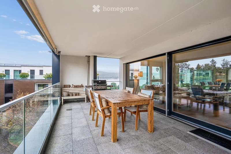 Modern balcony with a wooden dining table and chairs, a bench with baskets, and a grill, overlooking a scenic view.
