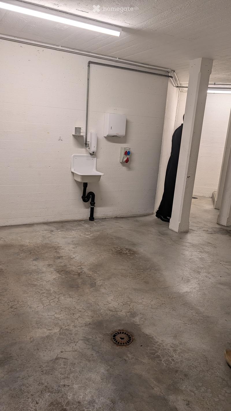Empty utility or storage room with a concrete floor, white walls, a sink, a water heater, and a drain in the center.