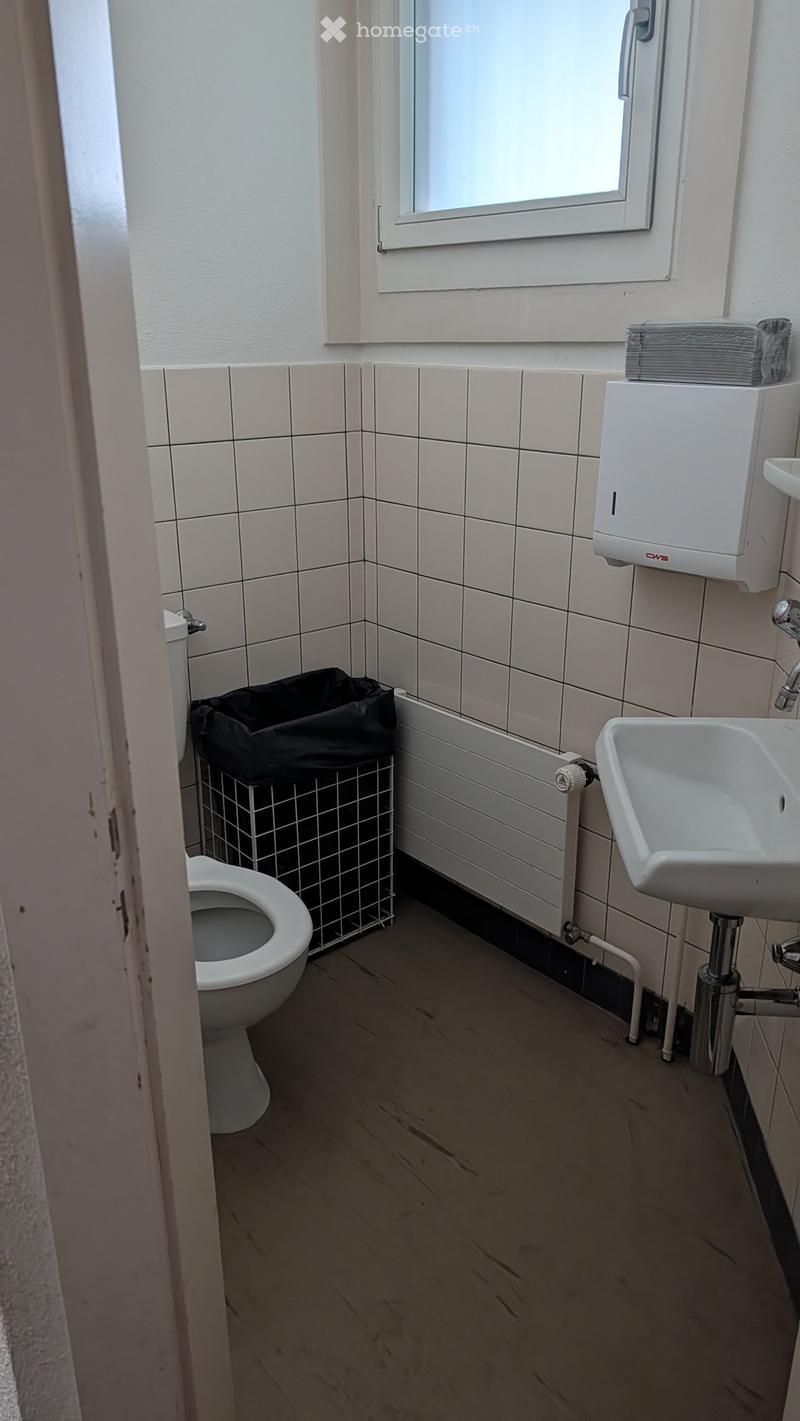 Small bathroom with white tiled walls, a toilet, a sink, a hand dryer, and a trash bin. The floor is wooden, and there is a window above the sink.