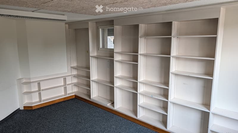 Empty room with built-in white shelving units and a small window, featuring a grey carpet and wooden baseboards.