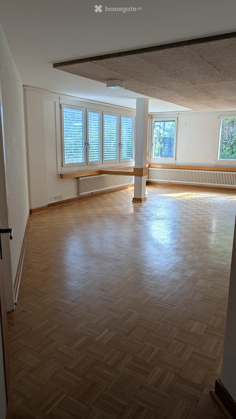 Empty room with wooden herringbone flooring, white walls, large windows with blinds, radiators, and a central column.