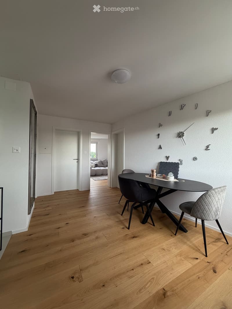 Furnished Room. Dining area with wooden floor, modern black dining table, chairs, and a wall-mounted clock.