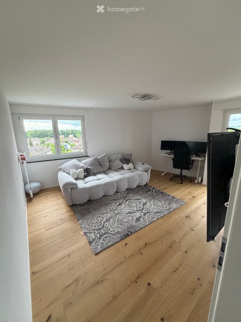 Furnished Room. A modern living room with a grey sectional sofa, a patterned rug, a desk with a computer and chair, and large windows offering a view of the neighborhood.