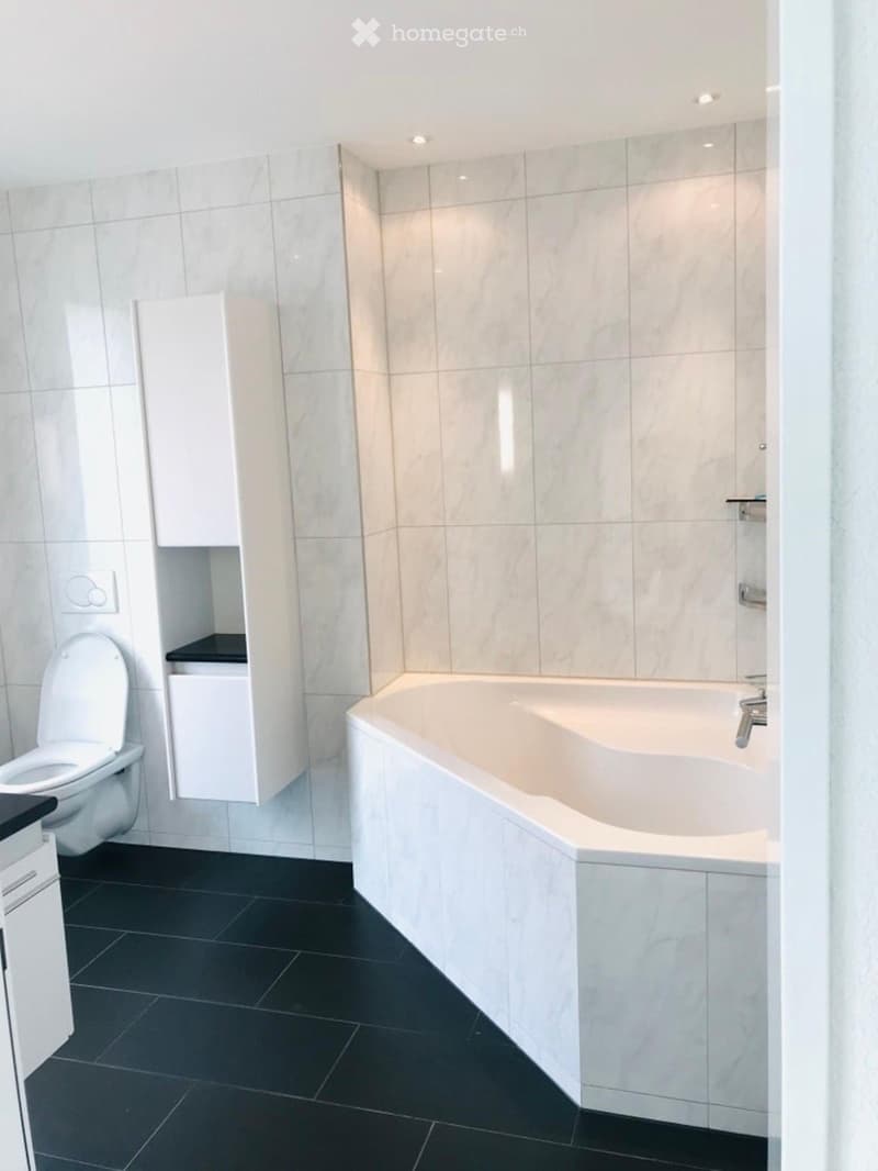 Modern bathroom with a white bathtub, toilet, and white cabinets. The walls are tiled in light marble, and the floor is black tile.