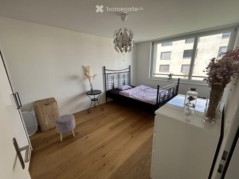 Cozy bedroom with wooden flooring, a metal-framed bed, a small round table, a wicker basket, a plush stool, and decorative vases on a nightstand.