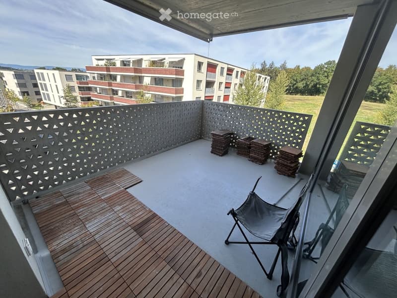 Balcony with wooden decking, grey folding chair, and stacks of wooden planks. Modern railing with geometric pattern.