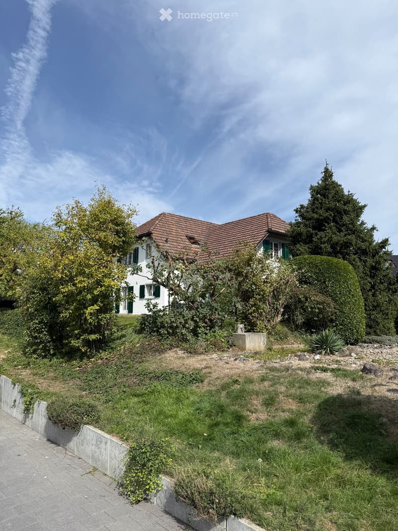 Two-story house with a red-tiled roof, white walls, green shutters, surrounded by trees and shrubs, with a raised garden area in front.