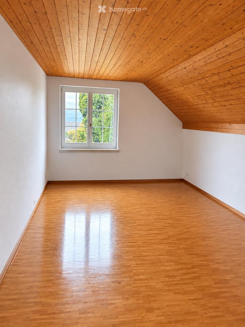 Empty room with wooden flooring and white walls, featuring a large window with a view of greenery outside.