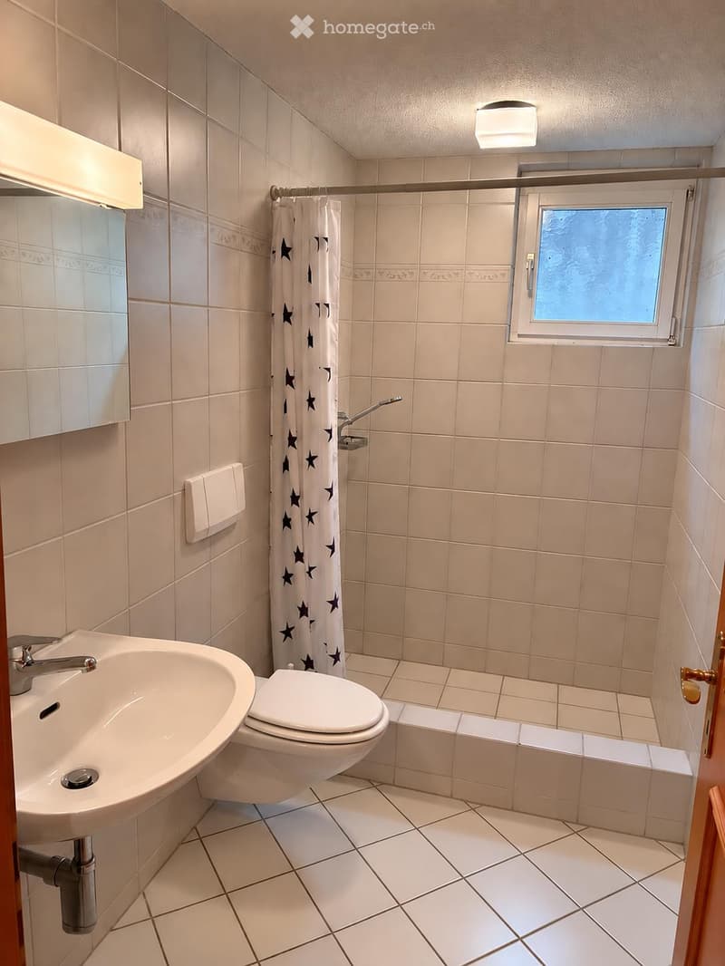 Bathroom with white tiled walls and floor, a sink, toilet, shower curtain with star pattern, and a small window.