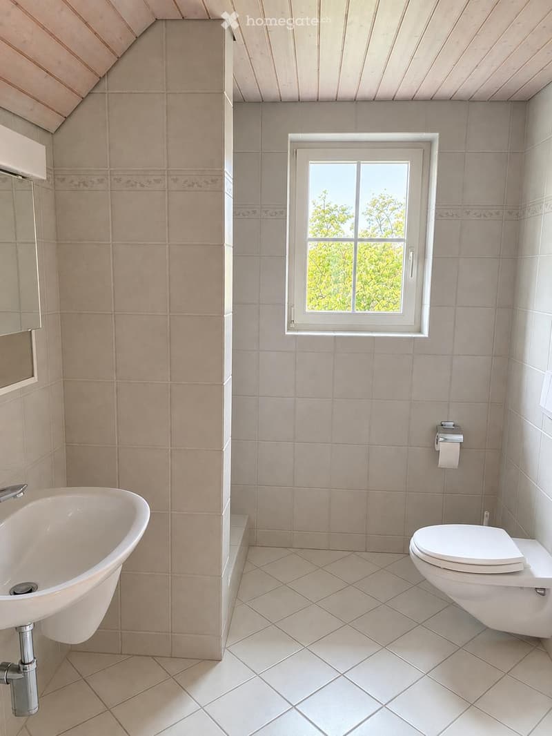 Bathroom with beige tiles, a bathtub, toilet, and a window with a view of greenery.