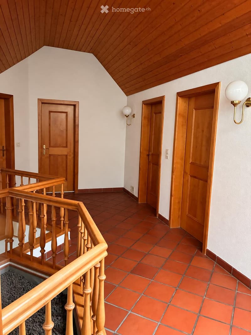 Wooden staircase with a wooden railing, red tiled floor, white walls, and several wooden doors leading to different rooms.