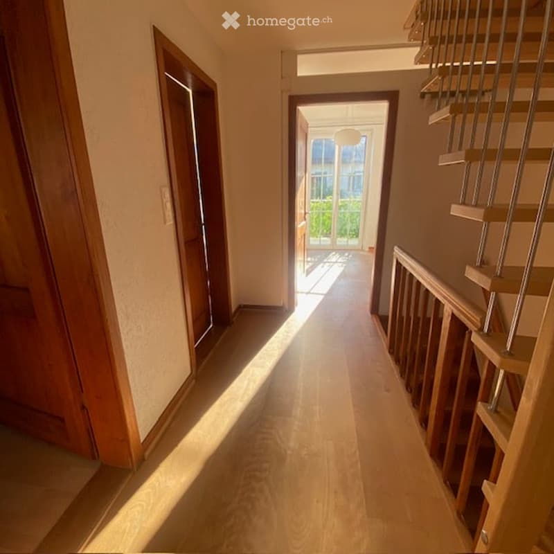 Bright hallway with wooden flooring, leading to a staircase and an open doorway to a room with large windows and a view of greenery.